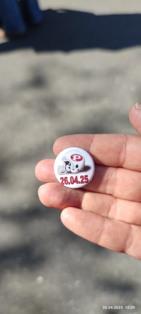 Ein kleiner Ansteckbutton mit einem Footballhelm, auf dem das Logo der Hamburg Pioneers abgebildet ist. Darunter ist das Datum der Auswärtsfahrt gedruckt, hier der 26.04.2025.